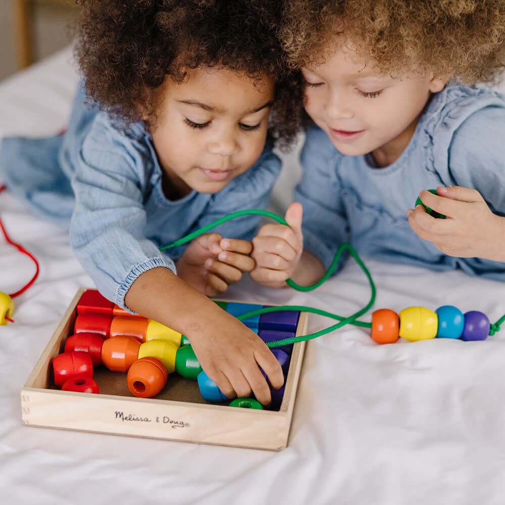 Two girls playing with the Melissa and Doug Primary Lacing Beads Set lacing the green lace with four beads on it